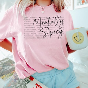 May include: A light pink t-shirt with the words "Mentally Spicy" in a black script font. The shirt also features a list of adjectives. A white mug with a yellow smiley face is held in the hand.