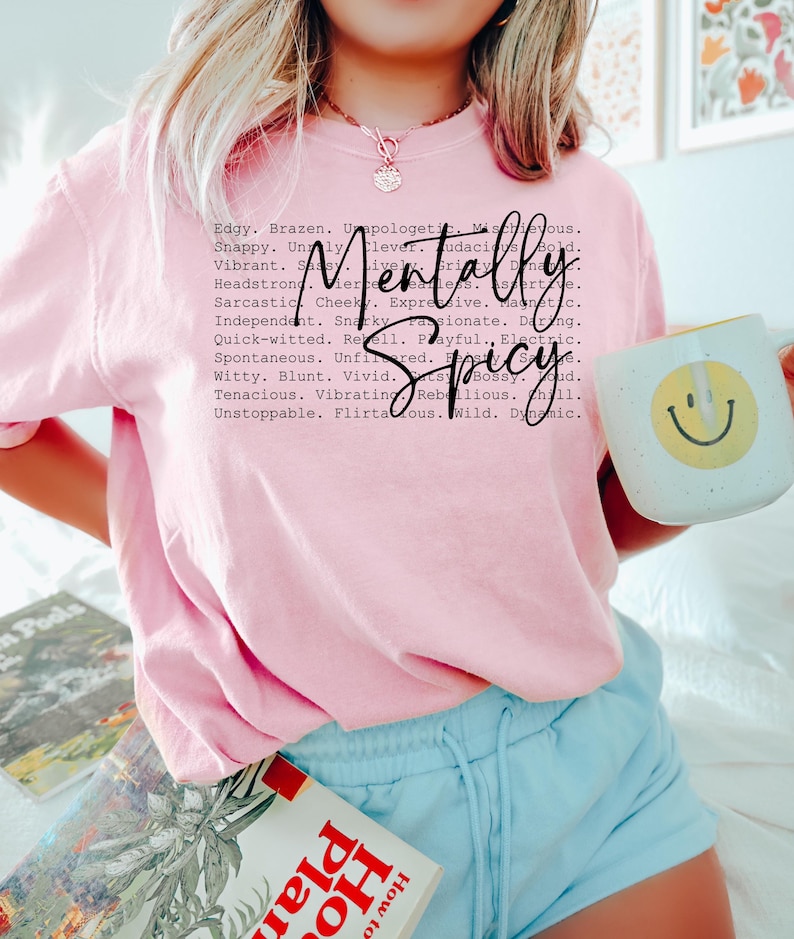 May include: A light pink t-shirt with the words "Mentally Spicy" in a black script font. The shirt also features a list of adjectives. A white mug with a yellow smiley face is held in the hand.