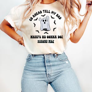Puede incluir: Camiseta color crema con un dise&ntilde;o de Halloween. El dise&ntilde;o presenta un fantasma de dibujos animados rodeado de murci&eacute;lagos, con el texto "GO AHEAD TELL MY DAD" y "WHAT'S HE GONNA DO? HAUNT ME?". La camiseta se combina con vaqueros azules.