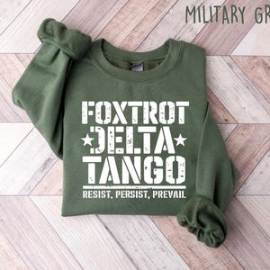 May include: Military green sweatshirt with the text "FOXTROT DELTA TANGO" in white, along with the words "RESIST, PERSIST, PREVAIL." The sweatshirt is folded, with the sleeves cuffed. The background is a light wood pattern.