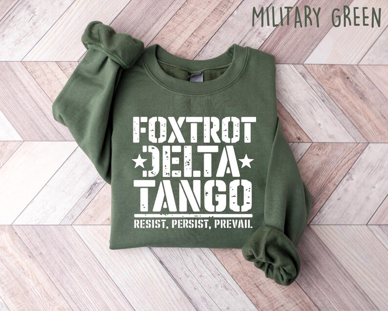 May include: Military green sweatshirt with the text "FOXTROT DELTA TANGO" in white, along with the words "RESIST, PERSIST, PREVAIL." The sweatshirt is folded, with the sleeves cuffed. The background is a light wood pattern.