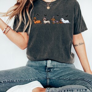 May include: Dark gray t-shirt featuring a Halloween-themed graphic of dachshunds in costumes. The dachshunds are dressed as a pumpkin, mummy, witch, and ghost. The word "BOO" is printed on the shirt. The shirt is paired with blue jeans.