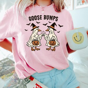 May include: A light pink sweatshirt with the words "GOOSE BUMPS" above a graphic of two cartoon geese dressed as ghosts, wearing witch hats, and holding jack-o'-lanterns. Bats are flying around the geese. A white mug with a yellow smiley face is held in the hand.