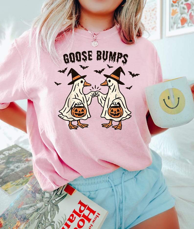 May include: A light pink sweatshirt with the words "GOOSE BUMPS" above a graphic of two cartoon geese dressed as ghosts, wearing witch hats, and holding jack-o'-lanterns. Bats are flying around the geese. A white mug with a yellow smiley face is held in the hand.