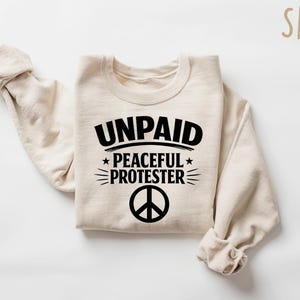 May include: A sand-colored sweatshirt with the words "UNPAID PEACEFUL PROTESTER" in black, along with a peace symbol. The sweatshirt is folded with the sleeves rolled up.