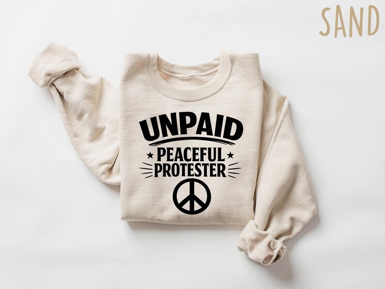 May include: A sand-colored sweatshirt with the words "UNPAID PEACEFUL PROTESTER" in black, along with a peace symbol. The sweatshirt is folded with the sleeves rolled up.