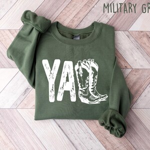 May include: A green sweatshirt with the word "Y'all" printed in white with a distressed look. The word is accompanied by a graphic of two white cowboy boots.