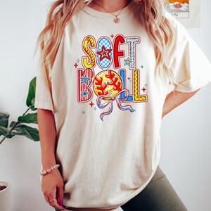 Puede incluir: Una camiseta de color crema con un dise&ntilde;o gr&aacute;fico colorido que dice "SOFTBALL" con una pelota de b&eacute;isbol y un lazo.