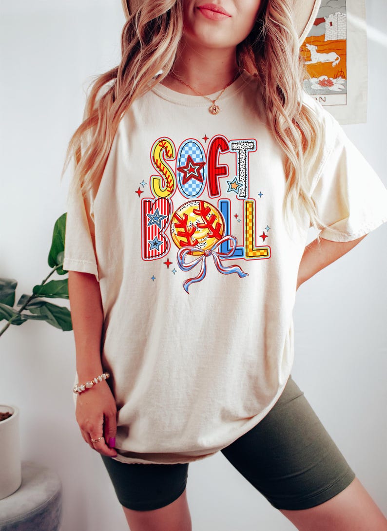 Puede incluir: Una camiseta de color crema con un dise&ntilde;o gr&aacute;fico colorido que dice "SOFTBALL" con una pelota de b&eacute;isbol y un lazo.