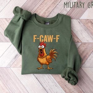 Puede incluir: Sudadera verde militar con el texto "F-CAW-F" y un gallo de dibujos animados con la boca abierta. El gallo es marr&oacute;n, naranja y rojo. La sudadera est&aacute; doblada sobre una superficie clara de vetas de madera.