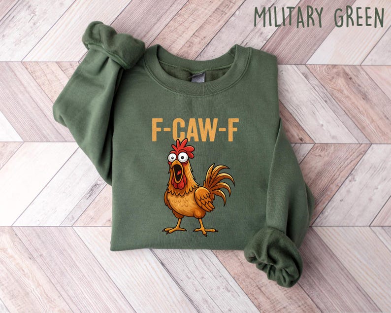 Puede incluir: Sudadera verde militar con el texto "F-CAW-F" y un gallo de dibujos animados con la boca abierta. El gallo es marr&oacute;n, naranja y rojo. La sudadera est&aacute; doblada sobre una superficie clara de vetas de madera.