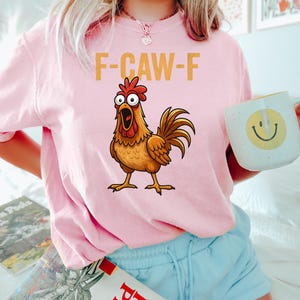 Puede incluir: Sudadera rosa claro con el texto "F-CAW-F" y una ilustraci&oacute;n de un gallo. El gallo es marr&oacute;n, con una cresta y barbillas rojas. La persona sostiene una taza blanca con una carita sonriente amarilla.