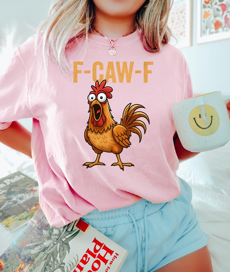 Puede incluir: Sudadera rosa claro con el texto "F-CAW-F" y una ilustraci&oacute;n de un gallo. El gallo es marr&oacute;n, con una cresta y barbillas rojas. La persona sostiene una taza blanca con una carita sonriente amarilla.