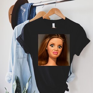May include: Black t-shirt featuring a doll's face with a surprised expression. The doll has brown hair, large eyes, and a puckered mouth. The shirt is hanging on a wooden hanger with other clothing items in the background.