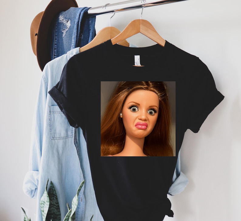 May include: Black t-shirt featuring a doll's face with a surprised expression. The doll has brown hair, large eyes, and a puckered mouth. The shirt is hanging on a wooden hanger with other clothing items in the background.