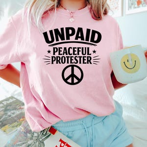 May include: A light pink sweatshirt with the words "UNPAID PEACEFUL PROTESTER" in black, along with a peace symbol. The person is holding a white mug with a yellow smiley face.