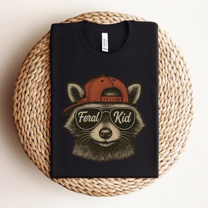 May include: Black t-shirt featuring a raccoon wearing a red cap and sunglasses. The raccoon has the words "Feral Kid" in a circular design on the front. The shirt is displayed on a woven, circular placemat.