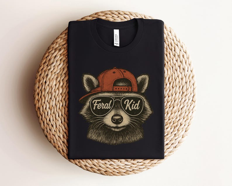 May include: Black t-shirt featuring a raccoon wearing a red cap and sunglasses. The raccoon has the words "Feral Kid" in a circular design on the front. The shirt is displayed on a woven, circular placemat.