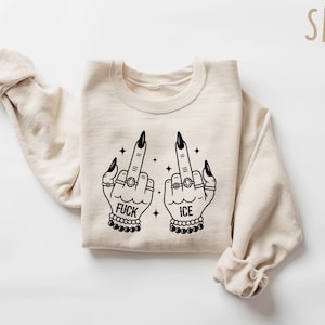 May include: Sand-colored sweatshirt with a black graphic of two hands giving the middle finger. The hands are adorned with rings, bracelets, and long black fingernails. The words "FUCK" and "ICE" are printed below the hands.