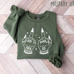 May include: Military green sweatshirt with white graphic of two hands giving the middle finger, adorned with rings and bracelets. The word "FUCK" is printed below one hand, and "ICE" below the other. The sweatshirt is folded on a light wood surface.
