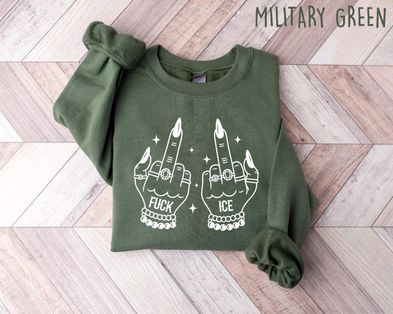 May include: Military green sweatshirt with white graphic of two hands giving the middle finger, adorned with rings and bracelets. The word "FUCK" is printed below one hand, and "ICE" below the other. The sweatshirt is folded on a light wood surface.