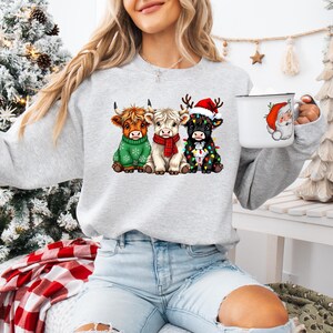 May include: A light gray sweatshirt featuring three Highland cows in festive holiday attire. One cow wears a green sweater, another a red scarf, and the third is adorned with Christmas lights and a Santa hat. A white mug with a Santa Claus graphic is also visible.