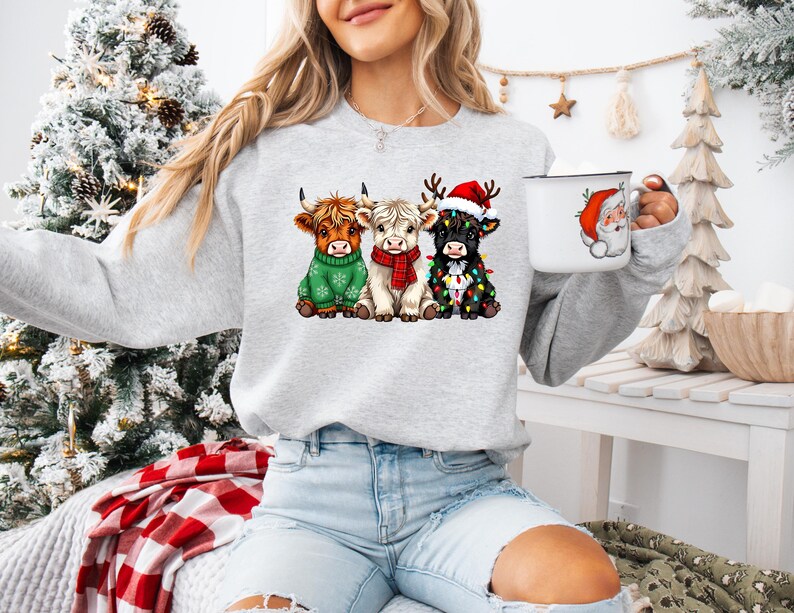 May include: A light gray sweatshirt featuring three Highland cows in festive holiday attire. One cow wears a green sweater, another a red scarf, and the third is adorned with Christmas lights and a Santa hat. A white mug with a Santa Claus graphic is also visible.