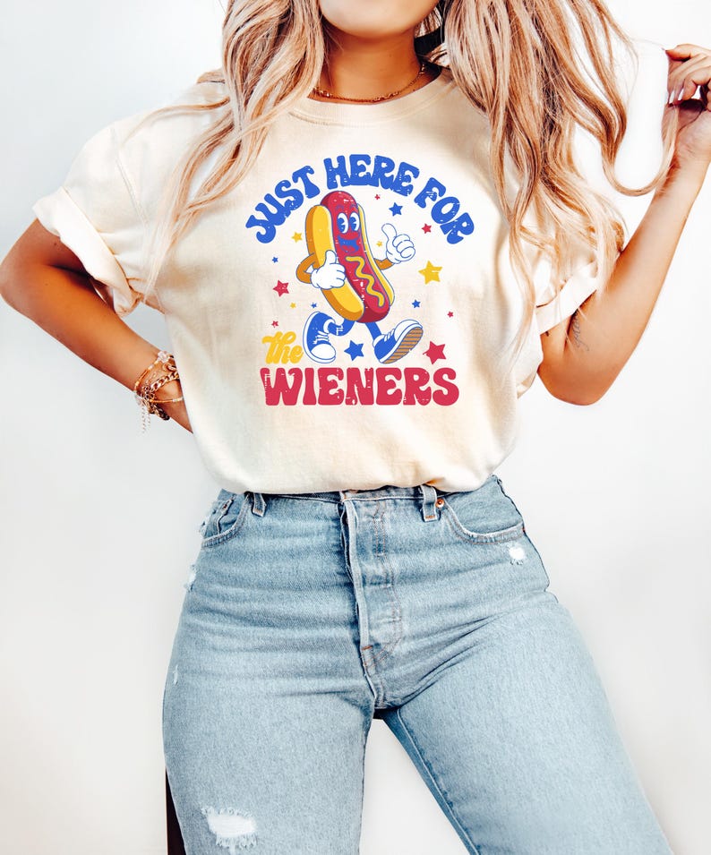 May include: Cream-colored t-shirt with the text "JUST HERE FOR the WIENERS" and a cartoon hot dog character. The hot dog has a red bun, yellow hot dog, and is giving a thumbs up. Blue jeans.