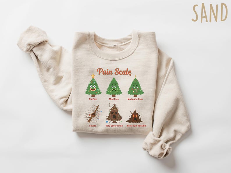 Puede incluir: Una sudadera de color arena con un gr&aacute;fico de una escala de dolor con &aacute;rboles de Navidad de dibujos animados. La escala va de "Sin dolor" a "Peor dolor posible". El texto "Pain Scale" est&aacute; arqueado sobre los &aacute;rboles.