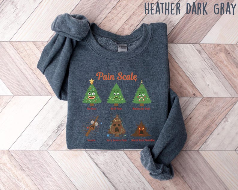 Puede incluir: Sudadera gris oscuro jaspeado con un gr&aacute;fico de "Escala de dolor" de &aacute;rboles de Navidad. El gr&aacute;fico presenta &aacute;rboles de Navidad de dibujos animados con diferentes expresiones faciales y niveles de dolor. El texto "Pain Scale" est&aacute; encima de los &aacute;rboles.