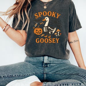 May include: Dark gray t-shirt with the words "SPOOKY GOOSEY" in orange, featuring a cartoon goose with a skeleton body, a jack-o'-lantern, and mushrooms. The shirt is worn with blue jeans and white shoes. Halloween themed apparel.