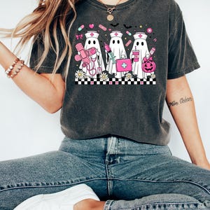 May include: Dark gray t-shirt featuring a graphic of three ghost nurses with medical supplies and a pumpkin. The design includes pink and white elements, such as a first aid kit, syringe, and pills. The shirt is paired with blue jeans and white sneakers.