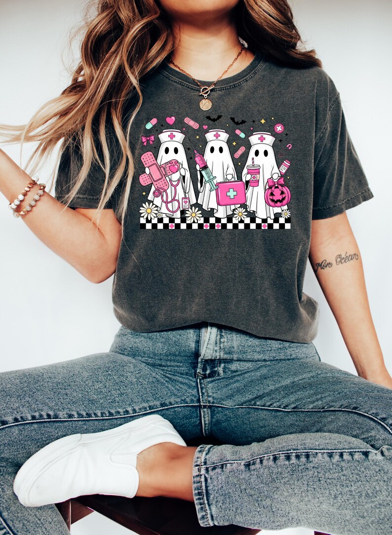 May include: Dark gray t-shirt featuring a graphic of three ghost nurses with medical supplies and a pumpkin. The design includes pink and white elements, such as a first aid kit, syringe, and pills. The shirt is paired with blue jeans and white sneakers.