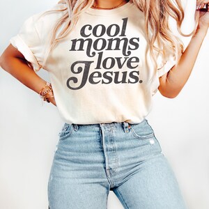 May include: Cream-colored t-shirt with the text "cool moms love Jesus" in a black, bold font. The shirt is paired with light-wash blue jeans. The sleeves of the shirt are rolled up. The overall style is casual and comfortable.