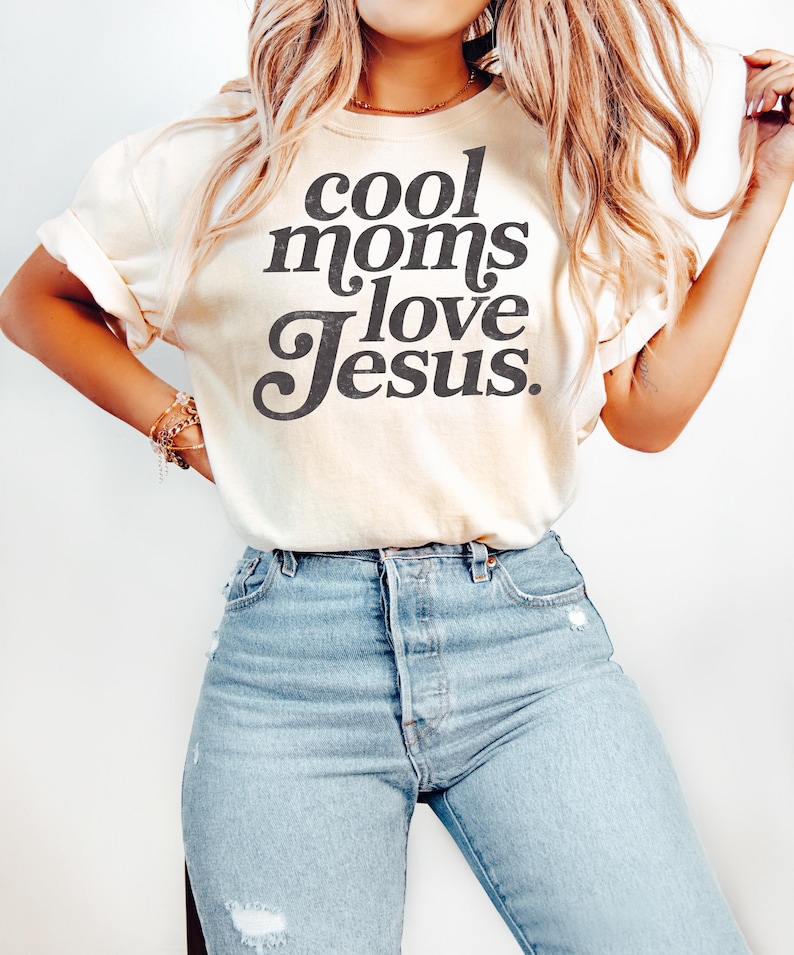 May include: Cream-colored t-shirt with the text "cool moms love Jesus" in a black, bold font. The shirt is paired with light-wash blue jeans. The sleeves of the shirt are rolled up. The overall style is casual and comfortable.