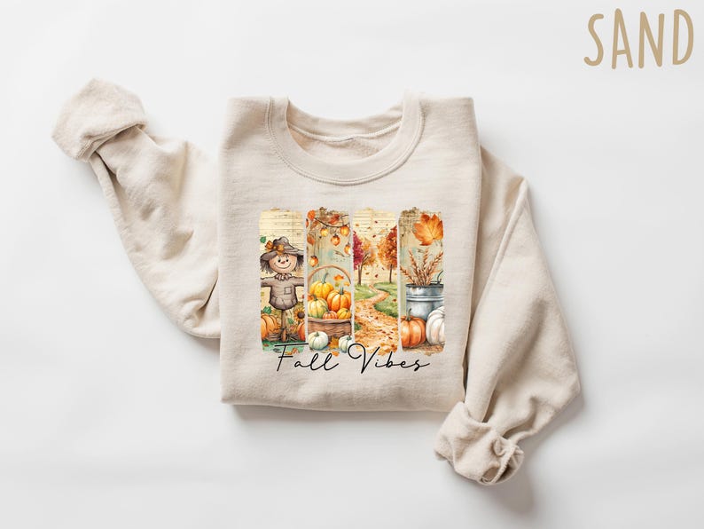 May include: Cream-colored sweatshirt with a watercolor design featuring a scarecrow, pumpkins, and fall foliage. The words "Fall Vibes" are written in a script font. The sweatshirt is folded with the sleeves draped.