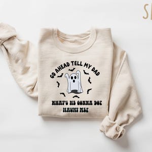 Puede incluir: Una sudadera de color arena con el texto negro "GO AHEAD TELL MY DAD WHAT'S HE GONNA DO? HAUNT ME?". Un fantasma de dibujos animados y murci&eacute;lagos rodean el texto. La sudadera est&aacute; doblada con las mangas enrolladas.
