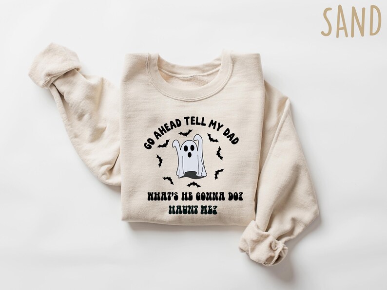 Puede incluir: Una sudadera de color arena con el texto negro "GO AHEAD TELL MY DAD WHAT'S HE GONNA DO? HAUNT ME?". Un fantasma de dibujos animados y murci&eacute;lagos rodean el texto. La sudadera est&aacute; doblada con las mangas enrolladas.