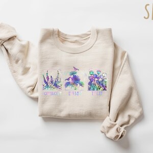 Op de afbeelding: Een beige sweatshirt met een bloemenontwerp en de woorden "To love, to trust, to life" gedrukt op de voorkant.