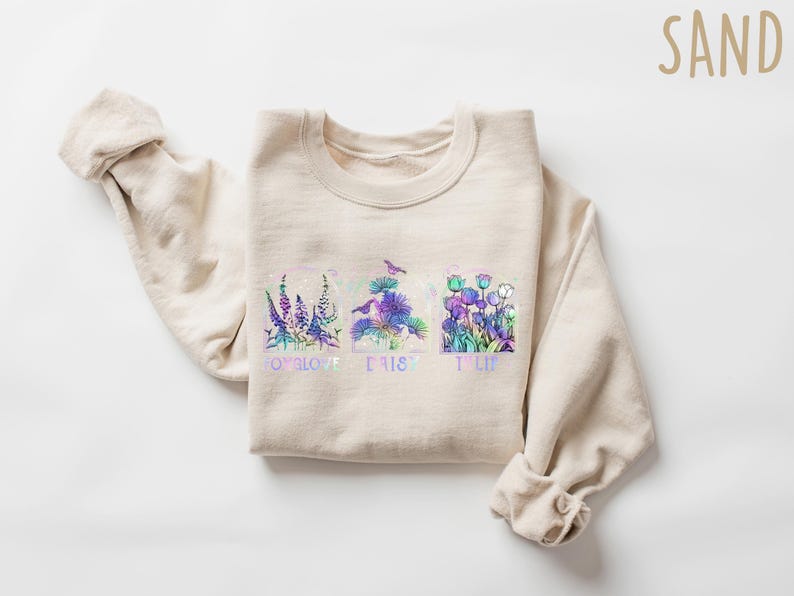 Op de afbeelding: Een beige sweatshirt met een bloemenontwerp en de woorden "To love, to trust, to life" gedrukt op de voorkant.