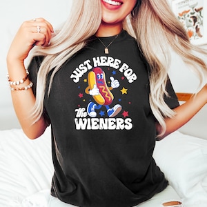 May include: Black t-shirt with the text "JUST HERE FOR THE WIENERS" and a cartoon hot dog character giving a thumbs up. The hot dog is wearing blue shoes and is surrounded by stars.