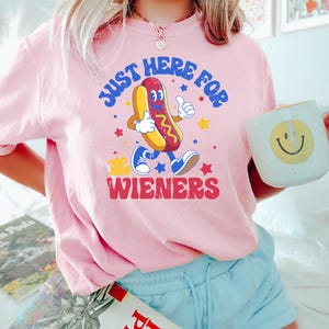 May include: Pink t-shirt with a retro cartoon hot dog graphic. The hot dog is walking and giving a thumbs up, with the text "JUST HERE FOR THE WIENERS" in a playful font. A white mug with a yellow smiley face is held in the hand.