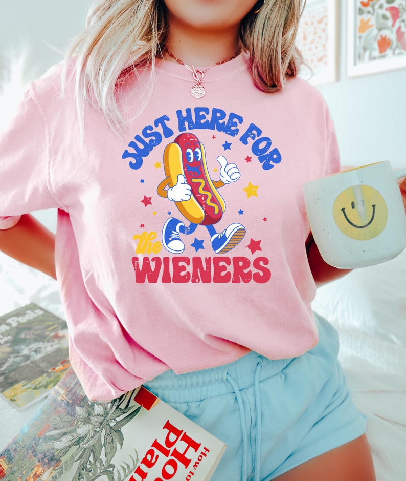 May include: Pink t-shirt with a retro cartoon hot dog graphic. The hot dog is walking and giving a thumbs up, with the text "JUST HERE FOR THE WIENERS" in a playful font. A white mug with a yellow smiley face is held in the hand.