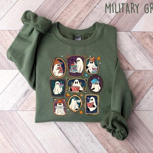 May include: Military green sweatshirt featuring a framed graphic of cartoon ghosts reading books. The ghosts are wearing hats and scarves. The sweatshirt is made of a soft material.