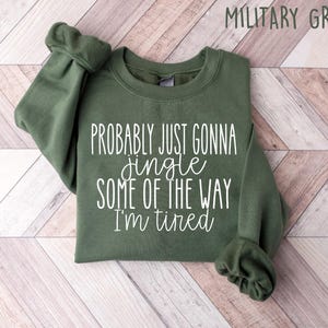 Op de afbeelding: Militairgroen sweatshirt met de tekst "Probably just gonna jingle some of the way I'm tired" in witte letters. Het sweatshirt is opgevouwen op een lichtbruin en wit chevron patroon oppervlak.