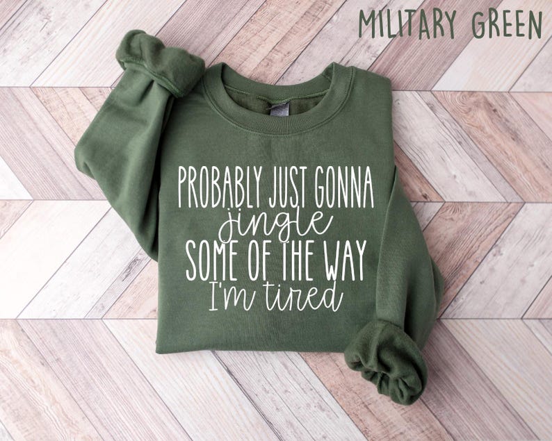 Op de afbeelding: Militairgroen sweatshirt met de tekst "Probably just gonna jingle some of the way I'm tired" in witte letters. Het sweatshirt is opgevouwen op een lichtbruin en wit chevron patroon oppervlak.