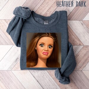 May include: Heather dark gray sweatshirt with a doll face graphic. The doll has wide eyes and a pursed expression. The sweatshirt is a solid color and has a crew neck.