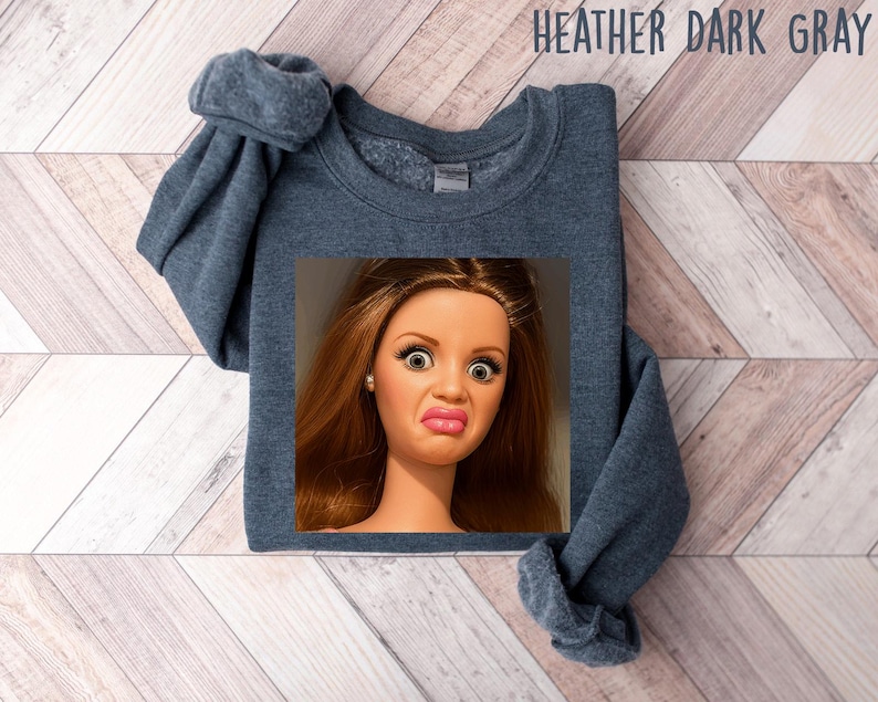 May include: Heather dark gray sweatshirt with a doll face graphic. The doll has wide eyes and a pursed expression. The sweatshirt is a solid color and has a crew neck.