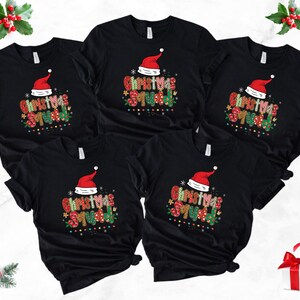 May include: Black t-shirts with a festive "Christmas Squad" design. Each shirt features a Santa hat and colorful text. The shirts are arranged on a white surface, with holiday decorations.