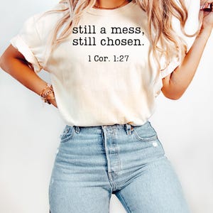 K&ouml;nnte beinhalten: Cremefarbenes T-Shirt mit dem Text "still a mess, still chosen." und dem Verweis "1 Cor. 1:27" in Schwarz. Das Shirt wird mit hellblauen Jeans kombiniert.